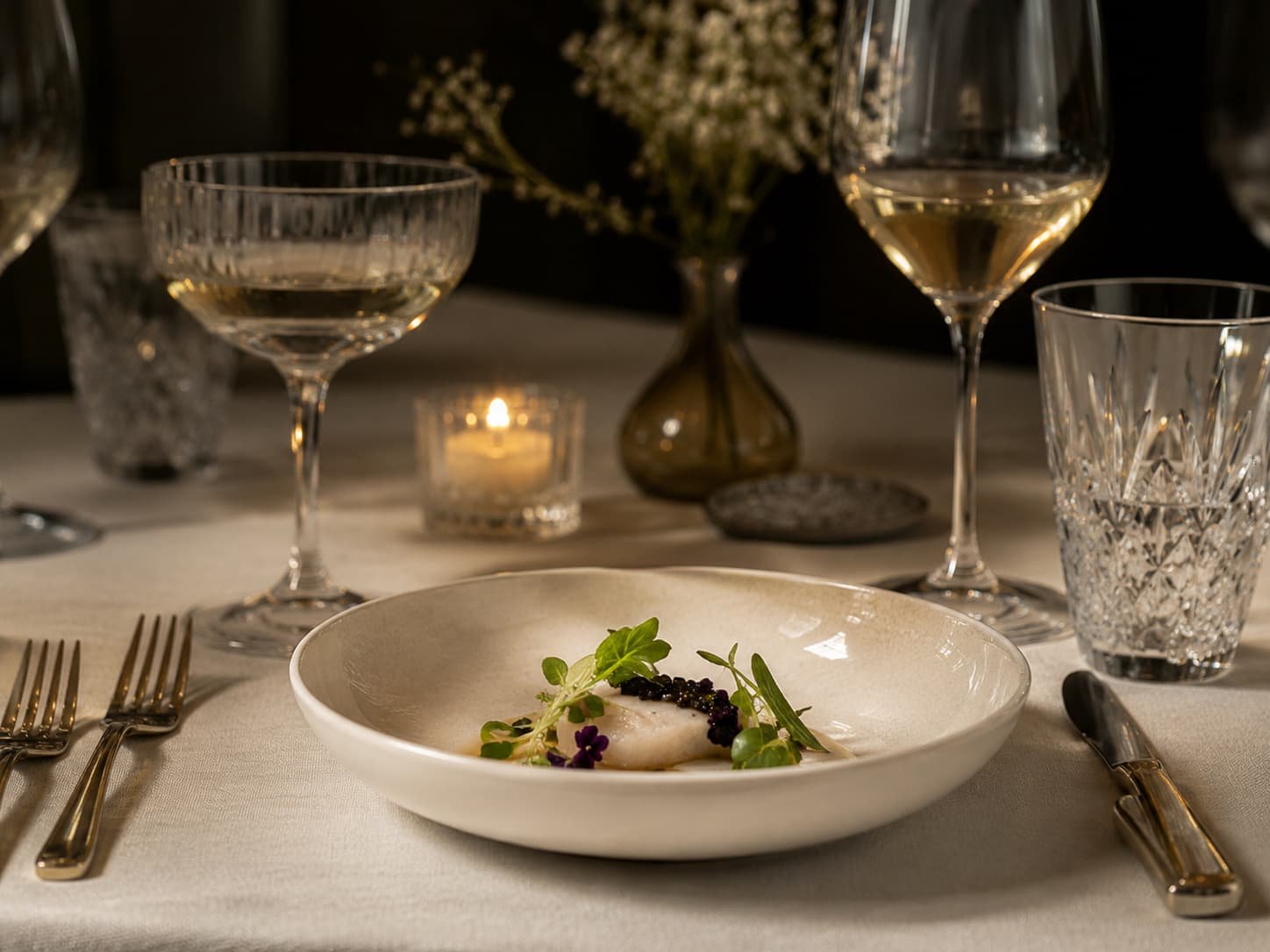 Elegantes Stillleben auf einem Fine-Dining-Tisch mit Glas, Besteck, Leinen, Kerzenlicht und perfekt angerichteter Speise.