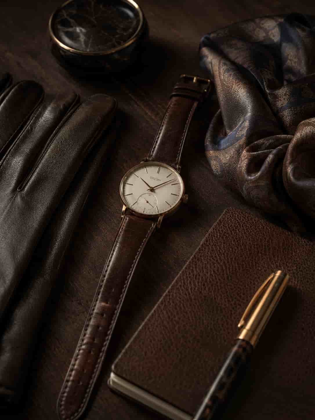 Zeitloses Accessoire-Stillleben mit schlichter Uhr, Lederhandschuhen, Notizbuch und Seidenschal auf dunklem Holz.