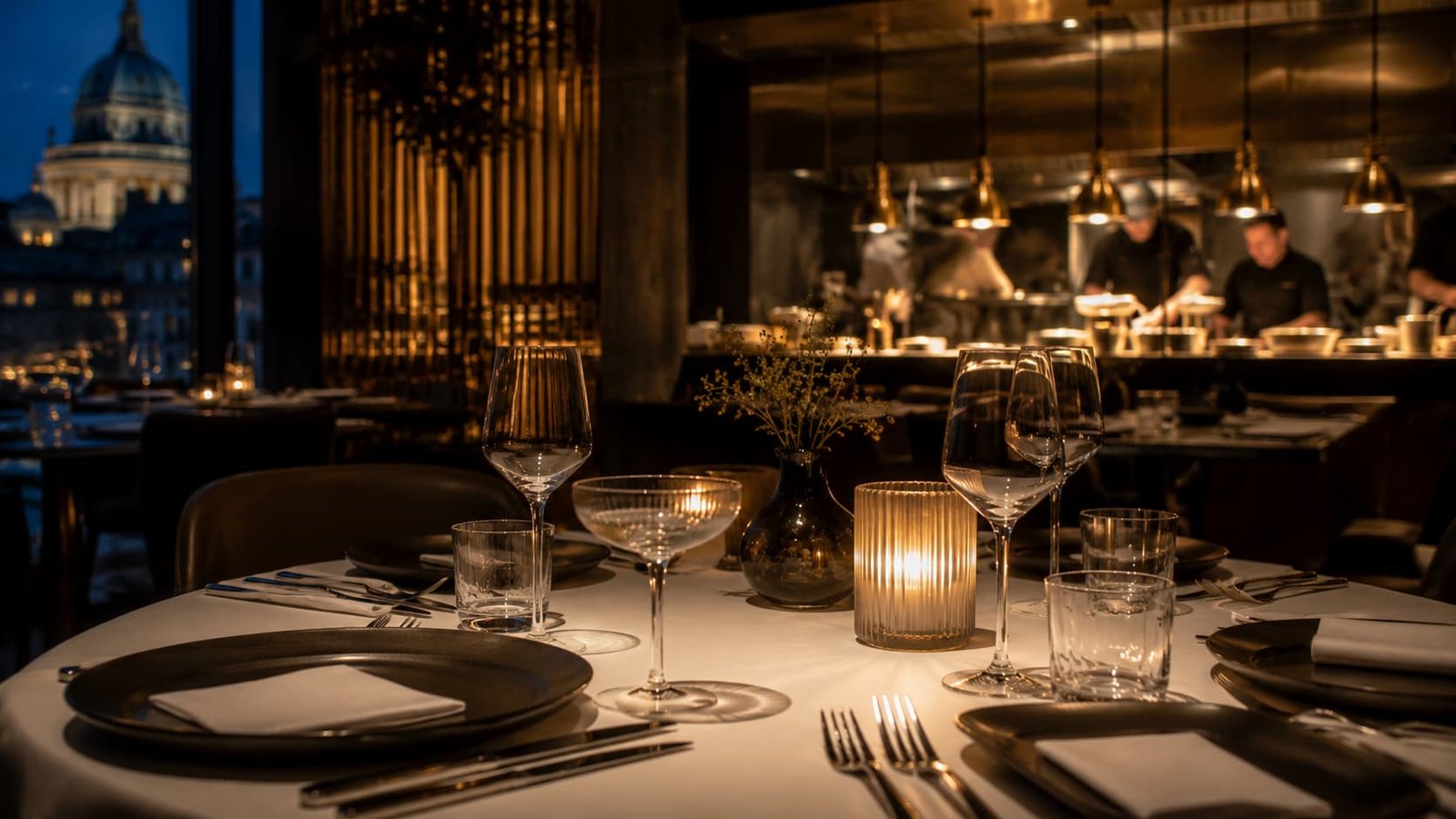 Fein gedeckter Restauranttisch mit Leinen, Keramik, Glas, Kerzenlicht und unscharfer offener Küche im Hintergrund.